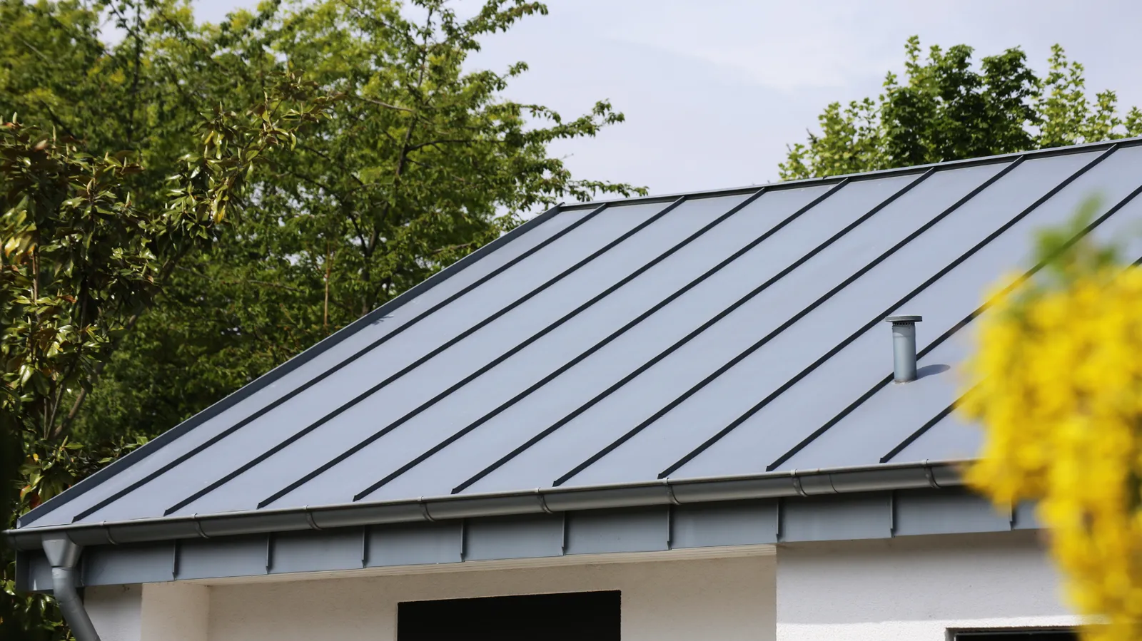 Standing seam metal roof edge with gutter beside leafy trees